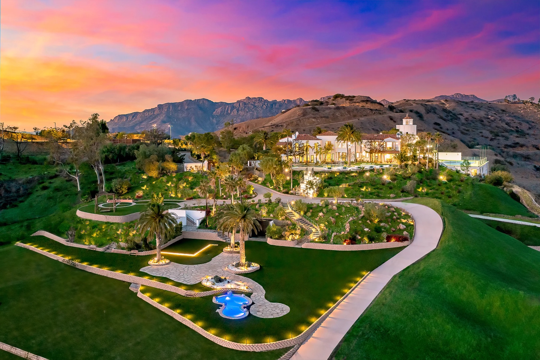 The Malibu estate at twilight: a white Italian villa with a bell tower, terraced gardens, palms, a lit pool, and the Santa Monica Mountains behind
