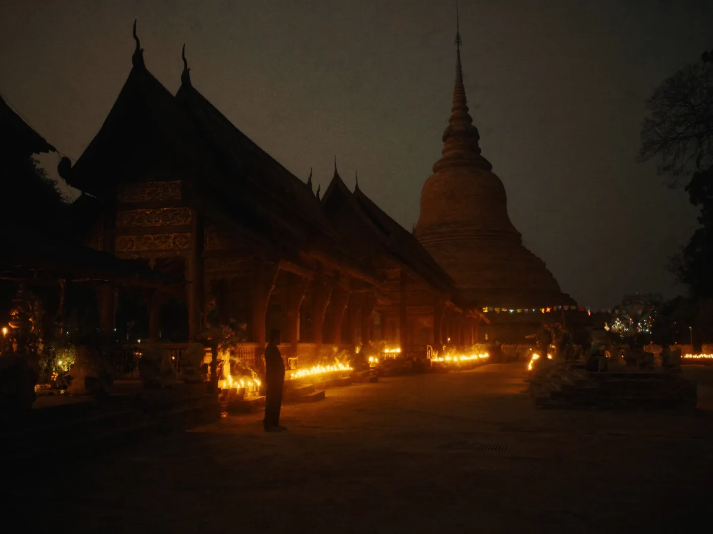 Candle offerings glowing beside a teak temple and ancient chedi at night